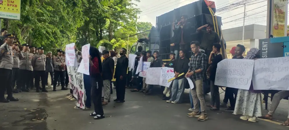 Massa aksi membawa poster tuntutan di pinggir jalan dengan sebuah mobil komando yang dilengkapi pengeras suara.