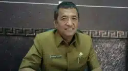 Foto potret Bambang Irianto mengenakan seragam dinas cokelat (ASN) dengan papan nama di dada, sedang duduk dan tersenyum ke arah kamera