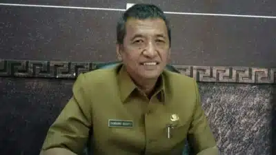 Foto potret Bambang Irianto mengenakan seragam dinas cokelat (ASN) dengan papan nama di dada, sedang duduk dan tersenyum ke arah kamera