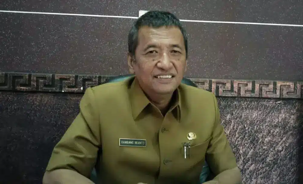 Foto potret Bambang Irianto mengenakan seragam dinas cokelat (ASN) dengan papan nama di dada, sedang duduk dan tersenyum ke arah kamera