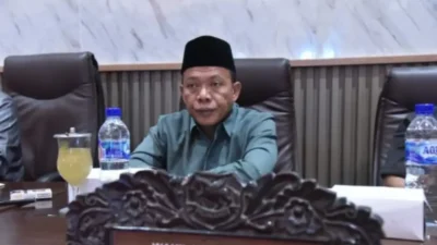 Wakil Ketua DPRD Sumenep, H. Dul Siam, mengenakan kemeja hijau dan peci hitam, duduk di meja rapat dalam sebuah pertemuan resmi.