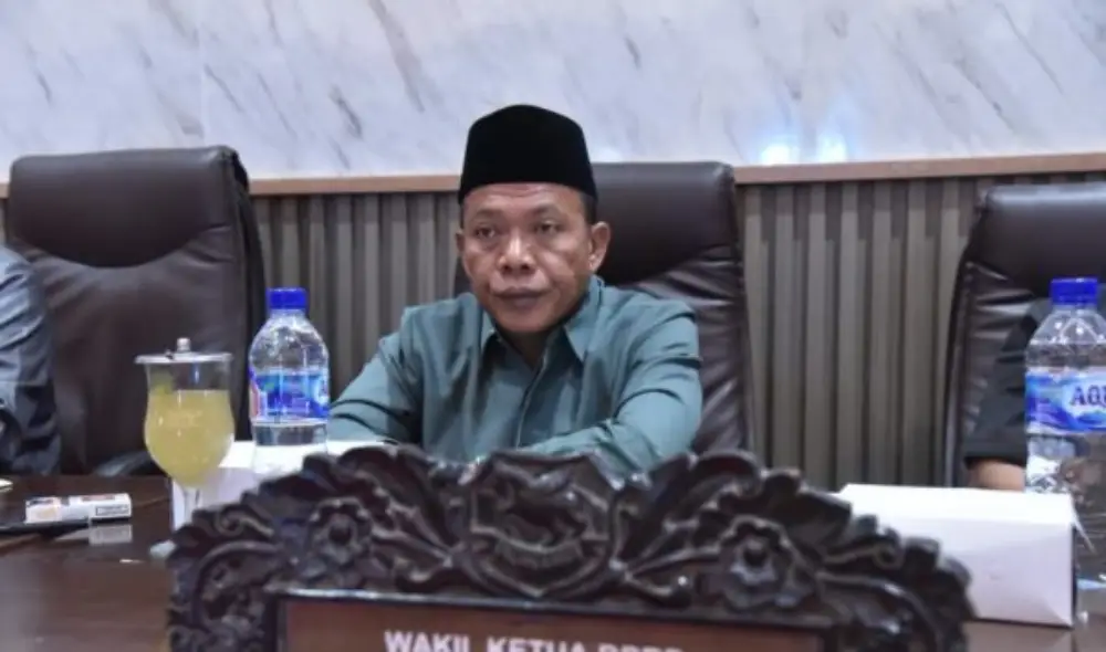 Wakil Ketua DPRD Sumenep, H. Dul Siam, mengenakan kemeja hijau dan peci hitam, duduk di meja rapat dalam sebuah pertemuan resmi.