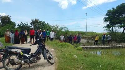 Sekelompok warga Desa Nambakor berdiri melakukan aksi protes di pinggir jalan dekat area tambak, dengan sebuah sepeda motor terparkir di latar depan dan jembatan bambu di latar belakang.