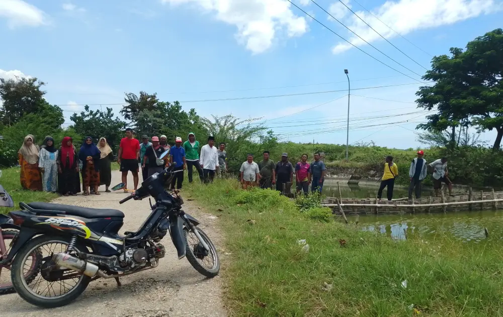 Sekelompok warga Desa Nambakor berdiri melakukan aksi protes di pinggir jalan dekat area tambak, dengan sebuah sepeda motor terparkir di latar depan dan jembatan bambu di latar belakang.