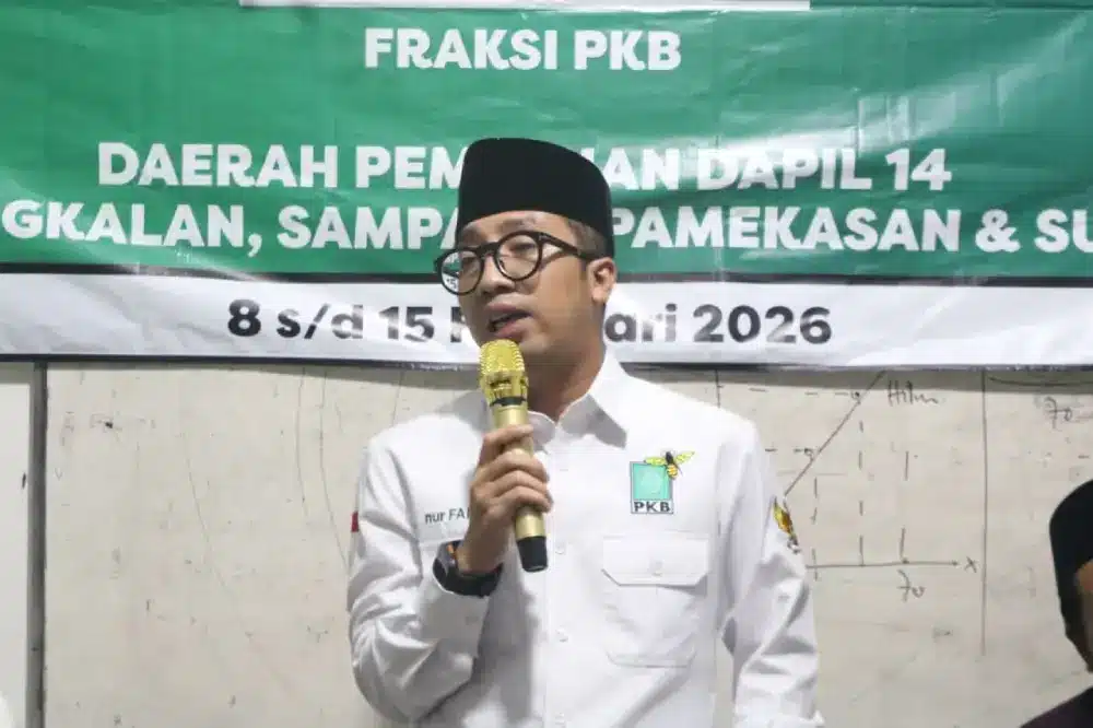 Nur Faizin, Anggota DPRD Jawa Timur dari Fraksi PKB, mengenakan kemeja putih berlogo PKB dan peci hitam, sedang berbicara menggunakan mikrofon emas dalam acara serap aspirasi di Saronggi, Sumenep. Di belakangnya terdapat spanduk hijau bertuliskan "FRAKSI PKB" dan latar belakang papan tulis dengan coretan grafik.