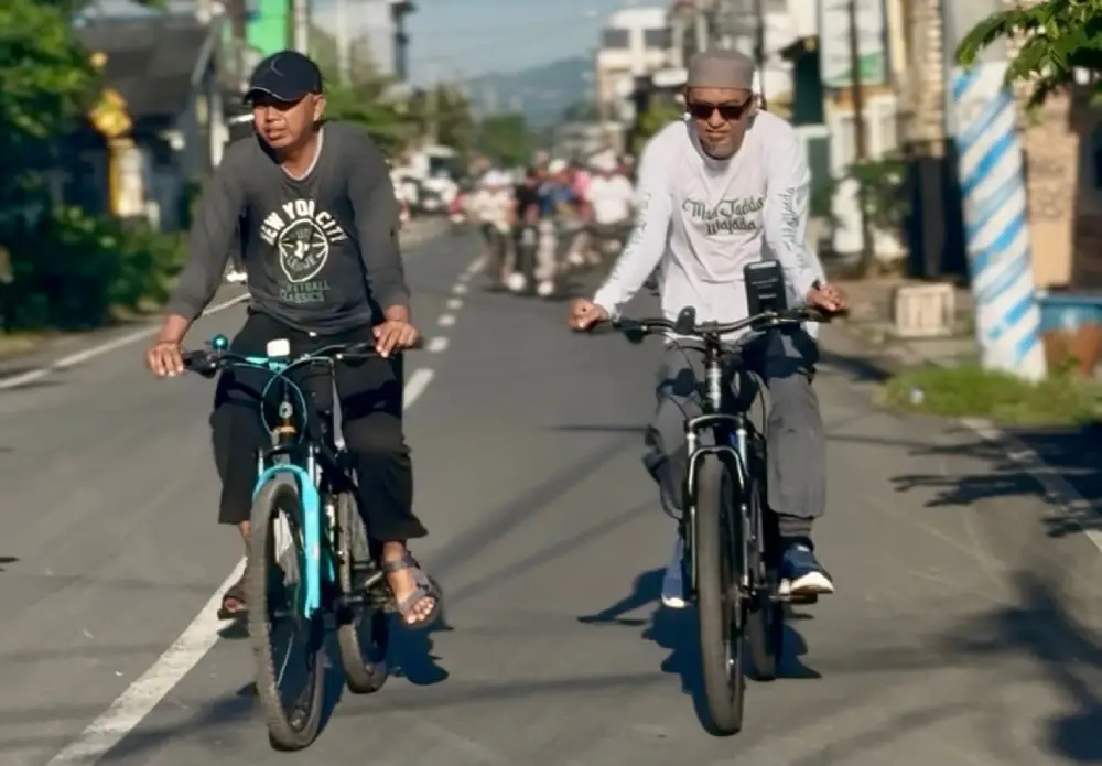 Dua orang pria, termasuk KH. Abuya Ach. Fadlan Masykuri, sedang mengendarai sepeda gunung di jalan raya saat acara Gowes Sehat Santri.