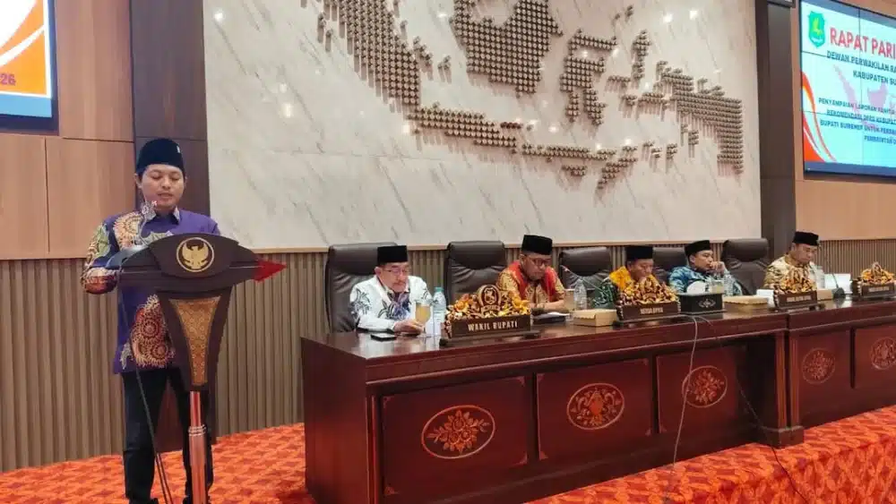 Suasana Rapat Paripurna DPRD Kabupaten Sumenep di ruang sidang utama. Seorang anggota DPRD berdiri di podium menyampaikan laporan, sementara di meja pimpinan duduk Wakil Bupati dan jajaran pimpinan DPRD dengan latar belakang dinding marmer berhiaskan dekorasi peta Indonesia.