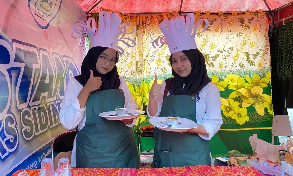 Dua orang siswi SMK SIDING PURI Lenteng mengenakan hijab, seragam putih, celemek hijau, dan topi koki unik dari kertas. Mereka berdiri di dalam stand pameran sambil membawa piring berisi produk "Sweet Potato Cream Brulee" dan memberikan pose jempol ke arah kamera.