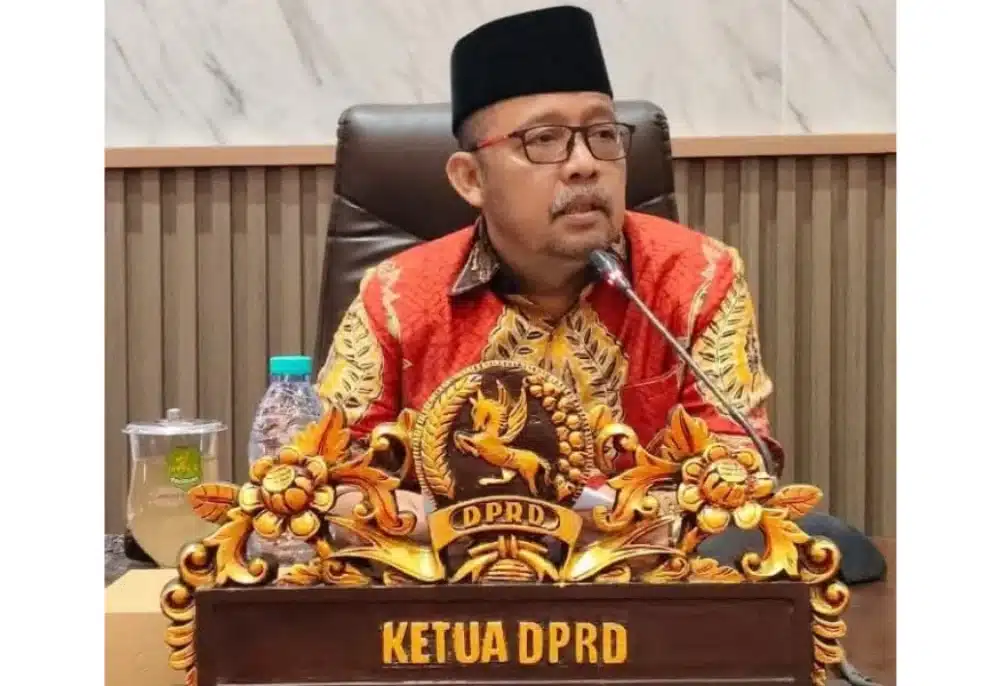 Ketua DPRD Sumenep, Zainal Arifin, mengenakan peci hitam dan batik merah, duduk di belakang meja pimpinan dengan papan nama "KETUA DPRD" saat membahas pengawasan program MBG.