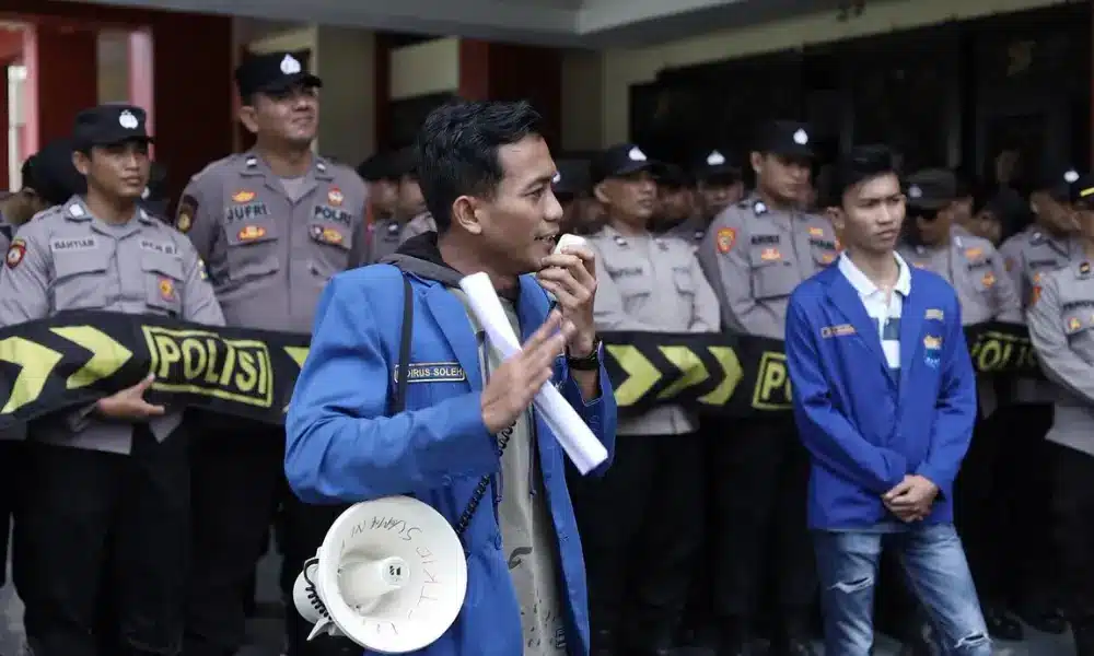 Khoirus Soleh, Ketua Umum PC PMII Sumenep, sedang berorasi menggunakan pengeras suara (megaphone) di depan barisan polisi yang berjaga di Kantor Pemkab Sumenep.