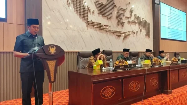 Sulahuddin, juru bicara pansus DPRD Sumenep, sedang berdiri di podium menyampaikan laporan pansus dalam Rapat Paripurna. Ia mengenakan seragam dinas hitam dan peci, sementara di latar belakang terlihat jajaran pimpinan termasuk Bupati dan pimpinan DPRD duduk di meja sidang dengan dekorasi peta Indonesia pada dinding