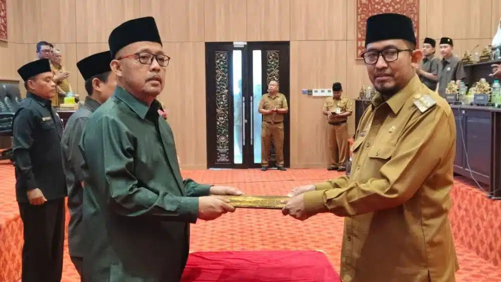 Ketua DPRD Sumenep sedang menyerahkan dokumen map kepada Bupati Sumenep dalam sebuah seremoni resmi di ruang rapat paripurna DPRD, disaksikan oleh sejumlah pejabat dan anggota DPRD lainnya.