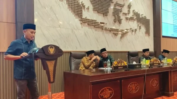 Irwan Hayat, juru bicara pansus DPRD Sumenep, sedang berdiri di podium menyampaikan laporan pandangan terkait Raperda Pengelolaan Pasar Rakyat dalam Rapat Paripurna. Ia mengenakan kemeja dinas biru tua dan peci hitam, dengan latar belakang jajaran pimpinan termasuk Bupati dan pimpinan DPRD yang duduk di meja sidang.