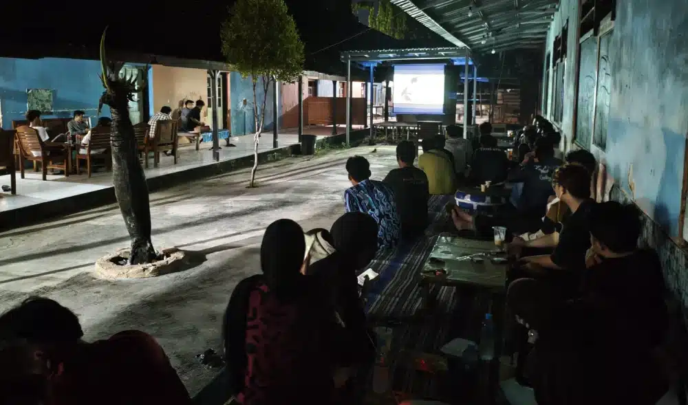 Ratusan mahasiswa duduk lesehan menghadap layar proyektor besar menyaksikan pemutaran film dokumenter Pesta Babi di area terbuka Kampoeng Kopi Sumenep saat malam hari.