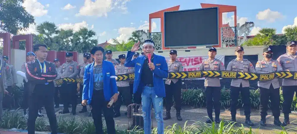 Foto aktivis mahasiswa PC PMII Sumenep sedang berorasi menggunakan mikrofon merah di depan barisan polisi yang memegang garis polisi (police line) dalam aksi unjuk rasa peringatan Hari Buruh.