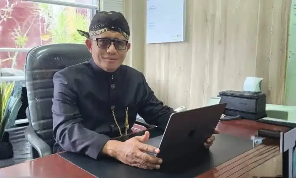 Dirut BPRS Bhakti Sumekar, Hairil Fajar, mengenakan pakaian adat dan blangkon, duduk menghadap laptop di meja kerjanya untuk memimpin program literasi keuangan syariah sekolah.