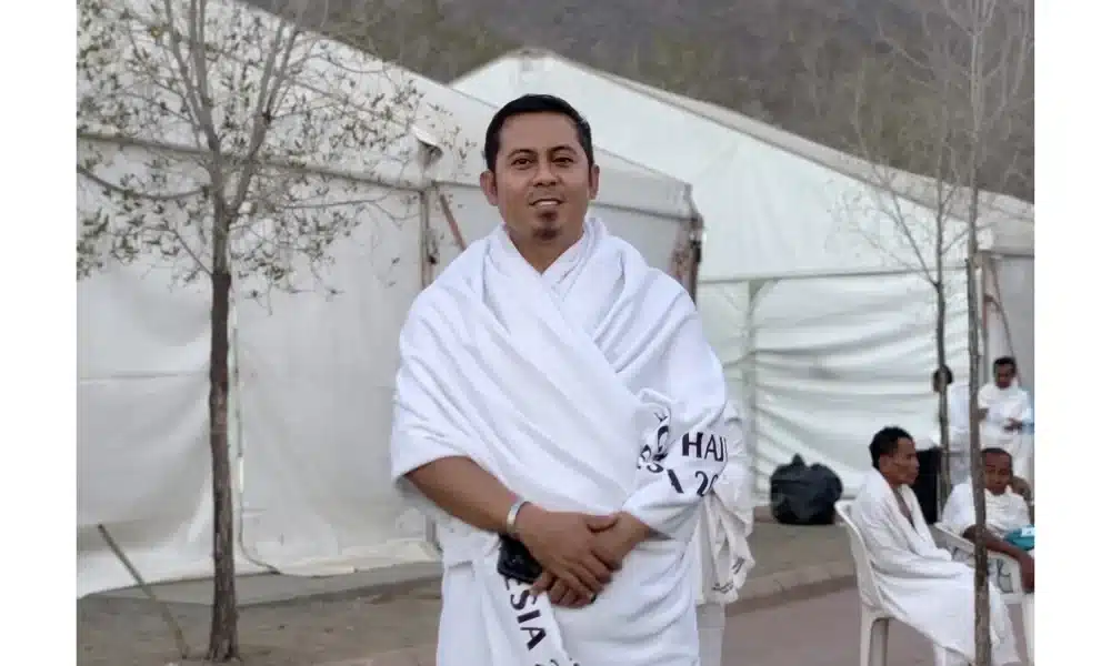 M. Hariri, jurnalis TV9 asal Sumenep, mengenakan pakaian ihram putih dengan tulisan "Haji Indonesia 2026", berdiri tersenyum di depan deretan tenda jamaah di Arafah atau Mina, Arab Saudi.