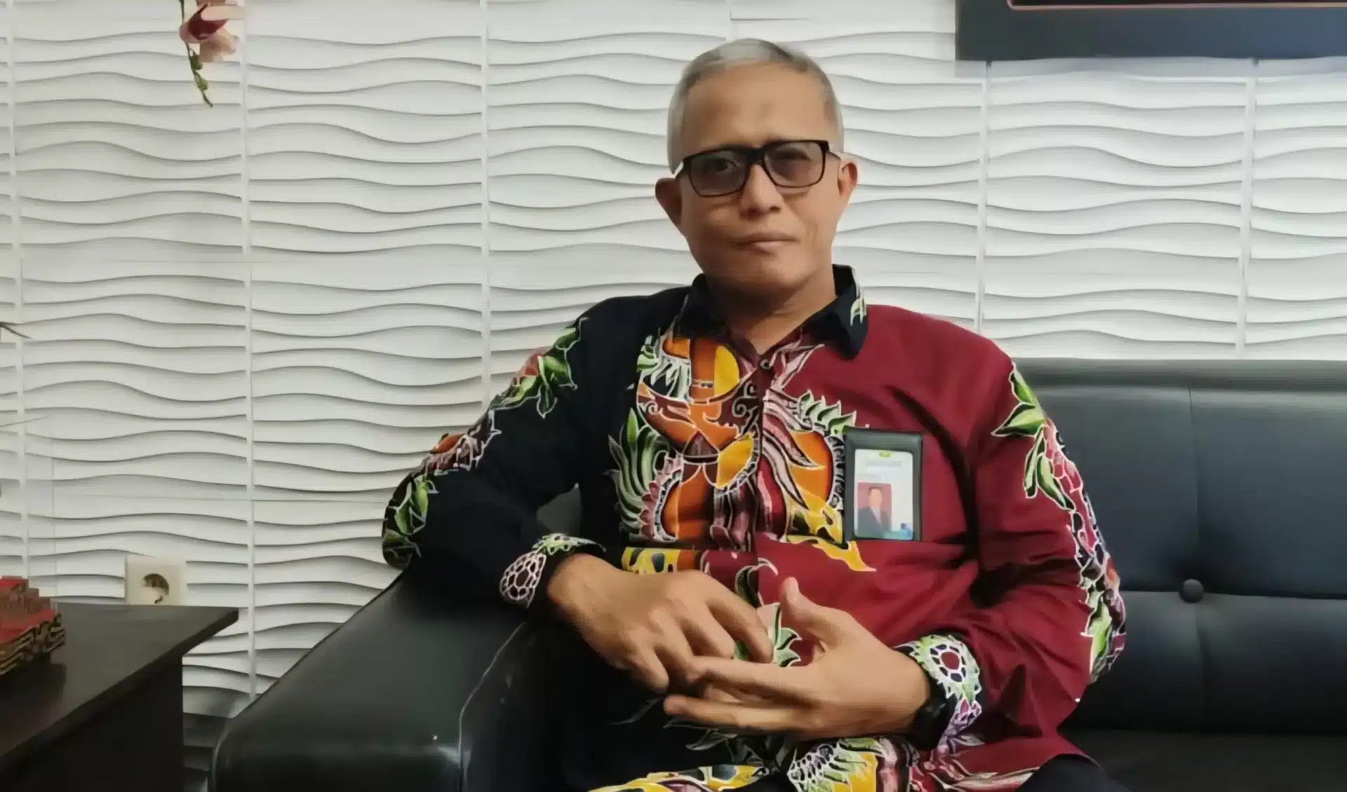 Direktur Utama BPRS Bhakti Sumekar, Hairil Fajar, mengenakan kemeja batik bermotif merah dan hitam serta kacamata, sedang duduk di sofa hitam dengan latar belakang dinding bermotif gelombang putih.