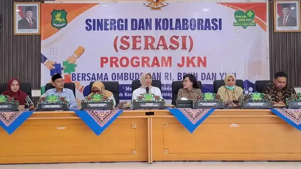 Suasana kegiatan Sinergi dan Kolaborasi (Serasi) Program JKN di Aula RSUDMA Sumenep. Tampak Direktur RSUD dr. H. Moh. Anwar Sumenep, dr. Erliyati, bersama perwakilan dari BPJS, Ombudsman RI, BPKN, YLKI, dan Dinkes P2KB duduk di meja depan dengan latar belakang spanduk besar bertuliskan "Sinergi dan Kolaborasi (Serasi) Program JKN".