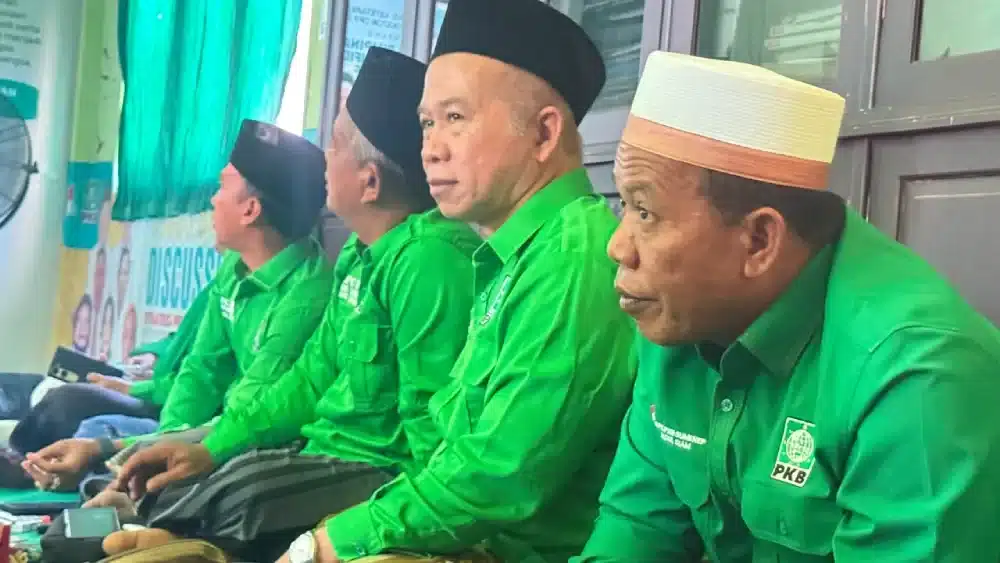 Foto KH Kamalil Ersyad mengenakan kemeja hijau seragam DPC PKB dan peci hitam, sedang duduk di depan mikrofon dalam suatu acara partai.