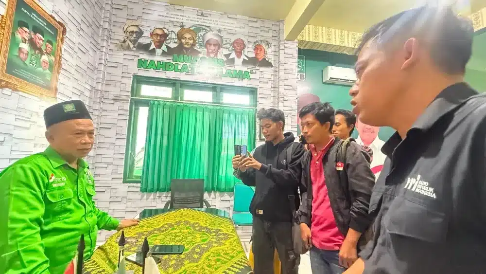 Alt: KH Kamalil Ersyad mengenakan kemeja hijau PKB dan peci hitam sedang duduk di meja kerja, didampingi oleh beberapa orang dan awak media di kantor DPC PKB Sumenep.