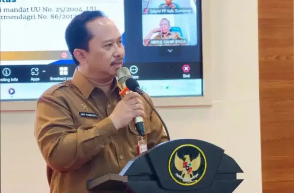 Foto Kepala Bappeda Sumenep Arif Firmanto mengenakan seragam dinas cokelat, sedang berbicara menggunakan mikrofon di podium hitam berlogo Garuda Pancasila dalam sebuah forum formal. Di latar belakang, tampak layar presentasi digital yang menampilkan teks regulasi dan jendela panggilan video daring dari peserta rapat.