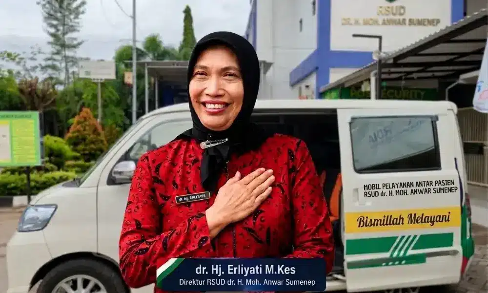 Foto dr. Hj. Erliyati M.Kes, Direktur RSUD dr. H. Moh. Anwar Sumenep, tersenyum ramah dengan tangan kanan di dada sebagai simbol pelayanan humanis berlandaskan Pancasila. Di latar belakang tampak mobil pelayanan antar pasien dan gedung rumah sakit