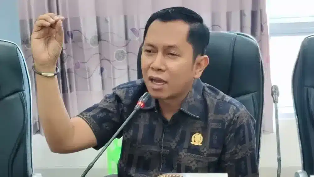 Foto Ketua Komisi IV DPRD SUMENEP, Mulyadi, mengenakan kemeja batik bermotif gelap dengan pin dinas di dada kiri, sedang berbicara memberikan pernyataan di depan mikrofon sambil mengangkat tangan kanannya.