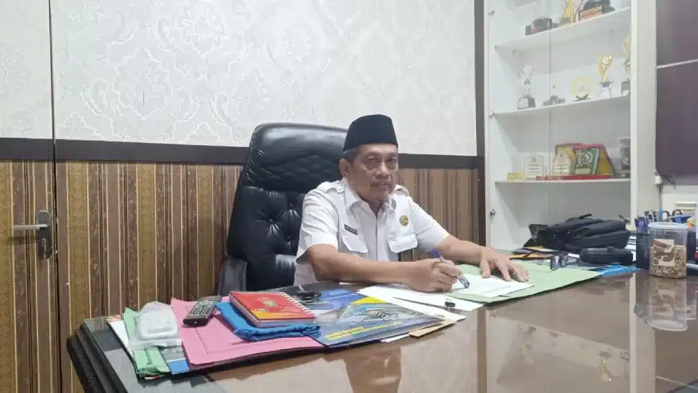 Kabid Bina Marga Dinas PUTR Sumenep, Slamet Supriyadi, mengenakan seragam dinas putih dan peci hitam, sedang duduk di meja kerjanya sambil memegang pena dan meninjau dokumen rencana proyek rekonstruksi jalan.