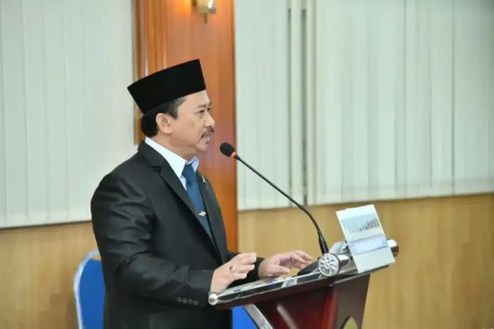 Kepala Bappeda Sumenep, Arif Firmanto, mengenakan setelan jas hitam dan peci, sedang berbicara di depan mikrofon pada podium acara kedinasan Pemkab Sumenep.
