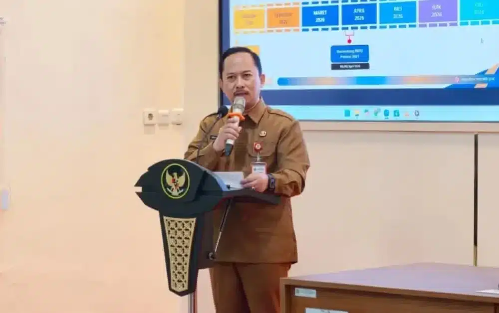 Kepala Bappeda Sumenep, Arif Firmanto, sedang memberikan sambutan di podium bersimbol Garuda dalam acara Pemkab Sumenep terkait penguatan reformasi birokrasi dan pelayanan publik, dengan latar belakang layar presentasi lini masa kebijakan daerah tahun 2026.