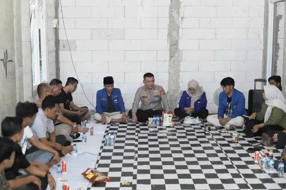 Sebuah foto yang diambil dari sudut pandang tinggi menunjukkan pertemuan formal antara sekitar belasan anggota PC PMII Sumenep dan jajaran Polresta Sumenep. Mereka duduk bersila dalam lingkaran besar di atas karpet bermotif catur di sebuah ruangan dengan dinding bata ekspos. Di tengah-tengah duduk Kapolresta Sumenep AKBP Henri Noveri Santoso yang mengenakan seragam dinas lengkap dan Ketua PC PMII Sumenep Khoirus Soleh yang mengenakan jas almamater PMII berwarna biru, diapit oleh anggota lainnya. Pertemuan berlangsung santai namun serius, dengan botol air mineral dan makanan ringan tersebar di depan mereka. Terdapat tanda air kecil bertuliskan 'HEADLINE NEWS' di sudut kanan bawah.