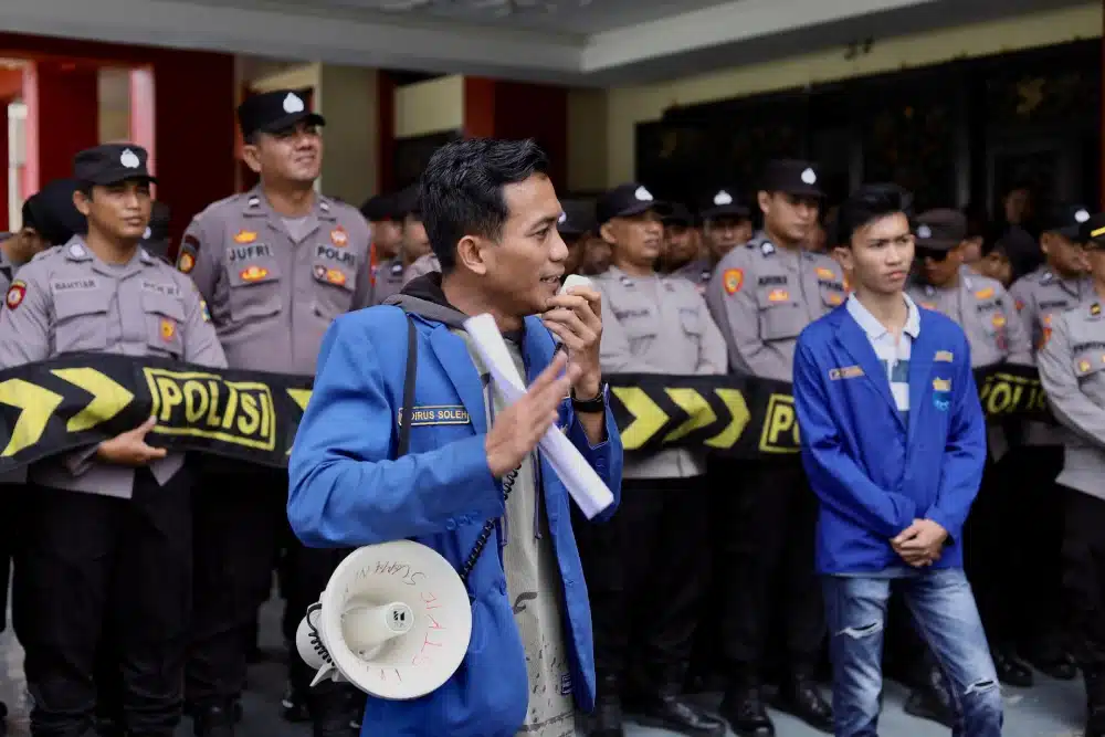 Seorang pria berkemeja dan almamater biru PC PMII (Khoirus Soleh) sedang berbicara menggunakan pengeras suara mikrofon di depan barisan aparat kepolisian yang bersiaga.
